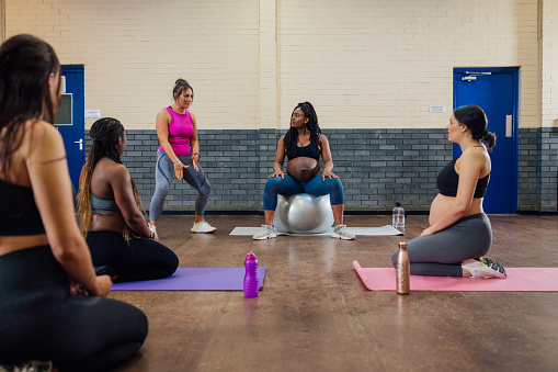 A group of pregnant mothers-to-be taking part in a yoga class in a community centre in Seaton Delaval, North East England. They are kneeling on yoga mats while watching one woman on an exercise ball at the front of the class, she is about to show the other women what they need to do for the exercise as the yoga instructor is talks them through it.