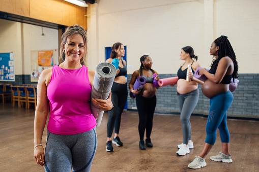 A group of pregnant mothers-to-be about to take part in a yoga class in a community centre in Seaton Delaval, North East England. The yoga instructor is standing close to the camera while looking at it and smiling as the rest of the women are standing in the background and talking.