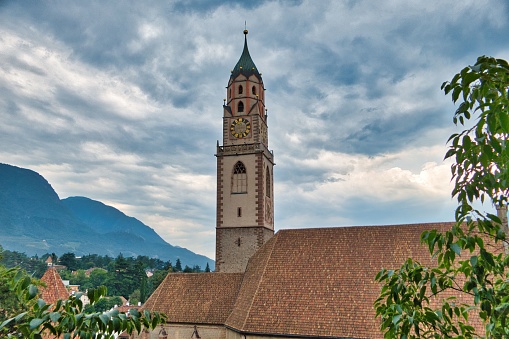 A picturesque scene featuring the clock tower of the City of Meran, South Tyrol, Italy, against the backdrop of lush green mountains