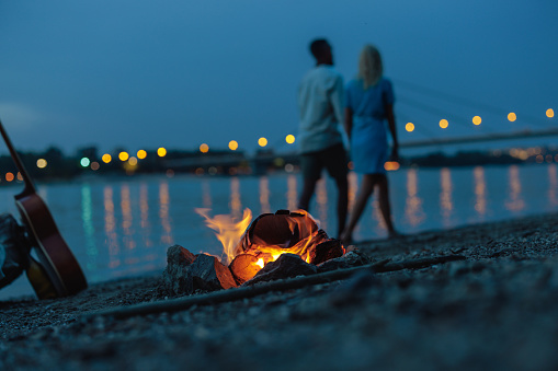 Shot of a bonfire by the river side with a guitar on the side and a couple walking on the shore