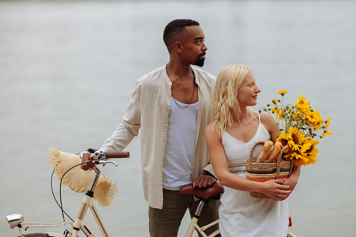 Beautiful and happy couple posing by the river while carrying sunflowers and a bike trying to find a perfect picnic spot