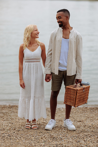 A lovely multiracial couple is holding hands while standing alongside the river, they are looking at each other and smiling