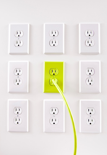 Wall of white electrical outlets in a row with center outlet green with a green cord plugged into it.