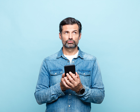 Thoughtful handsome mature man wearing denim shirt, holding smart phone, looking away. Studio shot.
