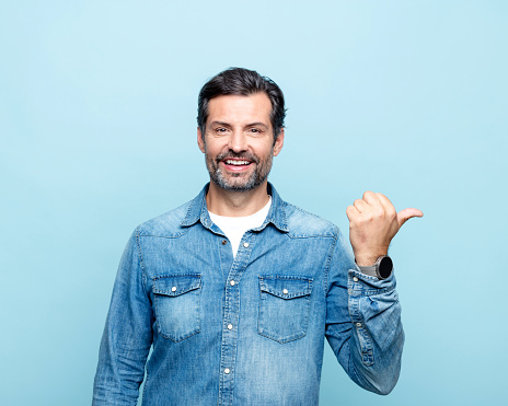 Happy mature man wearing denim shirt standing against blue background and pointing with thumb at copy space. Studio shot.