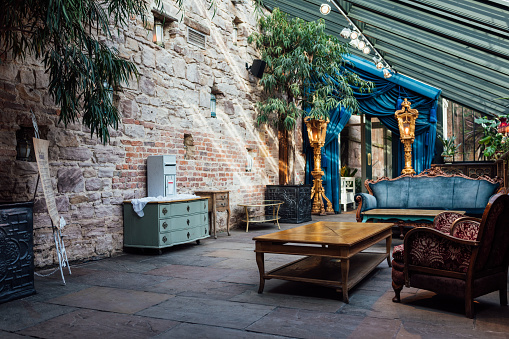 A shot of a spacious entryway at Newton Hall in Northumberland, North East England. It has a pitched glass roof which fills the space with natural light. There is a wall with rustic exposed stone and the floor is tiled. The furniture is an eclectic mix of decorative vintage pieces. Ornate freestanding lights stand either side of the door.