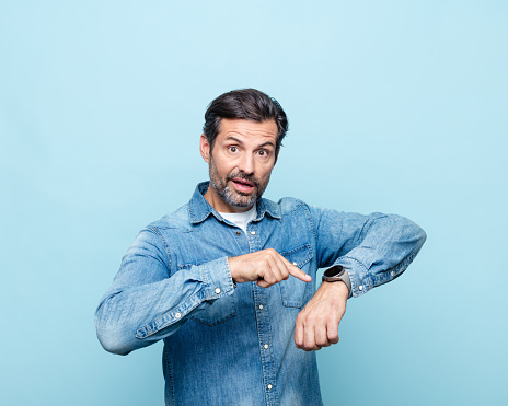 Surprised mature man wearing denim shirt pointing at watch with index finger, looking at camera. Studio shot, blue background.