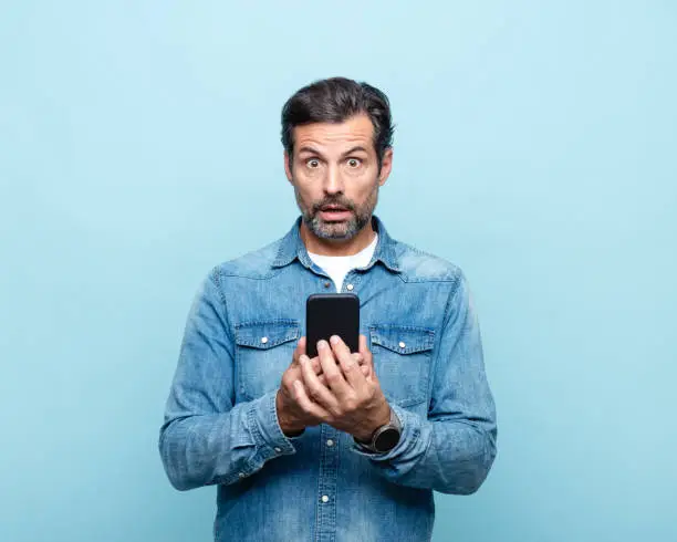 Shocked handsome mature man wearing denim shirt, holding smart phone, looking at camera. Studio shot.
