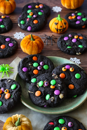 Stock photo showing close-up, elevated view of batch of Halloween white chocolate chip, black cocoa biscuits decorated with crunchy sugar coated, multicoloured chocolate sweets on a plate. Home baking concept photo.