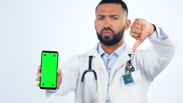 Phone, green screen and face of doctor with thumbs down hand gesture for healthcare in studio. Mockup, technology and portrait of male medical worker with disagreement expression by white background.