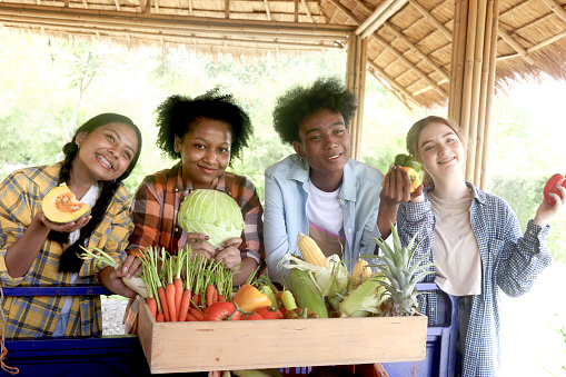 Group of happy Multiethnic teenager friend holding organic food products from their own farm at local market fair, smiling young diverse farmer selling fresh harvest fruit and vegetable at marketplace