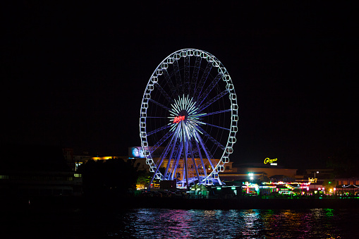 Illuminated ferris wheel of Asiatique The Riverfront in Bangkok seated in Bang Kho Laem at night seen from boat on Chao Praya river. Riverfront complex and promenade are offering many restaurants