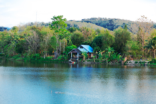 Small house idyllic at water and lake in Khao Yai. View over water. At right side Thai men and children are in scene. Children are playing at water. In background is tropical rainforest. Building has a blue roof