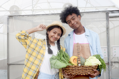 Portrait of two multiethnic teenager friend, Asian girl and African boy holding fresh harvest fruit vegetable basket at front of vegetable farm greenhouse, young gardener working in agricultural field