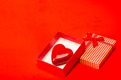 View of red gift box with ribbon an object in heart shape over red color background isolated.IMAGE MADE IN STUDIO AS CONCEPTUAL IMAGE FOR VALENTINE'S DAY