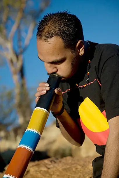 Mid shot of a young Aboriginal man playing a colourful didgeridoo in the outback of Australia.