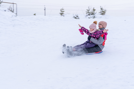 Two happy mother and daughter skating down a snowy hill on a sled. Two sisters in winter clothes have a rest in winter. Winter outdoor recreation. Family winter holidays