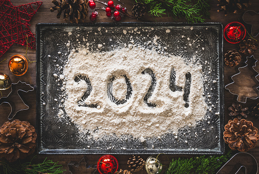 A baking tray with a pile of flour with the number 2024 written on it, surrounded by Christmas decorations and cookie cutters. Happy new year 2024.