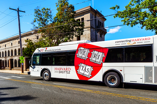 Boston, Massachusetts, USA - October 13, 2023: A whIte Harvard University Shuttle Bus on North Harvard Street going past Harvard Statium. The Harvard red shield with the VERITAS motto is on the side of the bus. The Harvard Shuttle buses serve the Cambridge and Allston (Boston) campuses.
