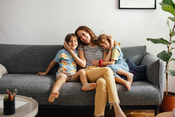 A Happy Beautiful Woman Keeping Her Eyes Closed While Enjoying Spending Her Leisure Time At Home With Her Cute Children A smiling Caucasian female hugging her adorable daughter and son while sitting on the sofa. boy and girl looking at camera stock pictures, royalty-free photos & images