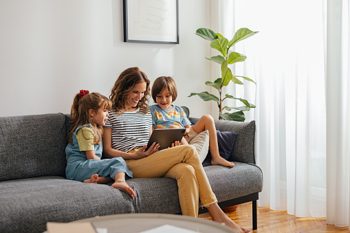 A wide angle view of a smiling Caucasian female and her adorable daughter and son using their tablet while sitting on the sofa in the living room.