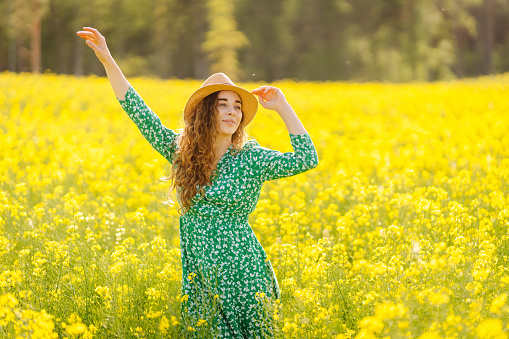 Happy woman is having fun and walking in a blooming rapeseed field. A beautiful woman in a bright dress and hat enjoys the sunny weather. Concept of nature, relaxation.
