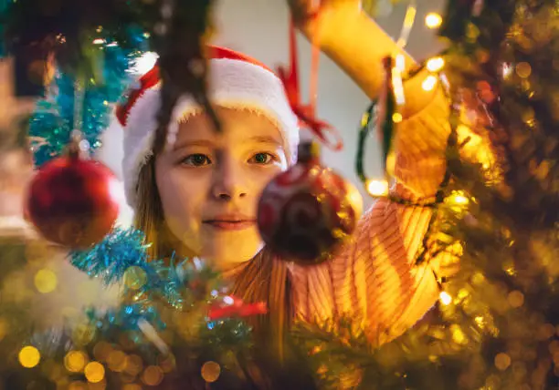 Cute little girl in red Santa hat hanging a magic tree decoration ball on the Christmas fir-tree on fairy tale x-mass wishes night. Charming childhood and winter holidays concept inside branches image