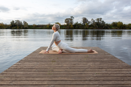 Mature woman practicing yoga, doing exercises on four limbs, push-ups from the floor, Half pose of the royal pigeon, Eka Pada Rajakapotasana, training, dressed in sports clothes. outdoor nature background. Autumn evening by the river.