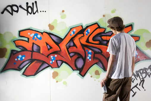 A teenager graffiti artist spray painting a wall and creating street art associated with rebelling and vandalizing in youth culture.