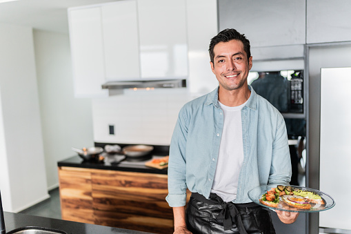 Portrait of a mid adult man holding a plate with toasted bread and guacamole in the kitchen at home