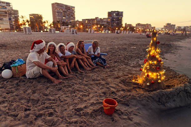 Multi generation family are having fun during summer Christmas.  They are enjoying evening on the beach with Christmas tree. They are sitting near the tree, talking and laughing.Canon R5