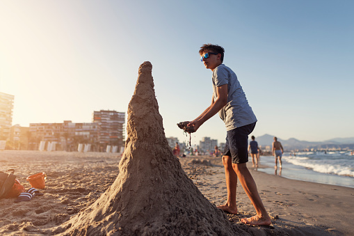 Teenage boy having fun during summer Christmas.  The boy is building a sand Christmas tree made on a summer day evening.\nCanon R5