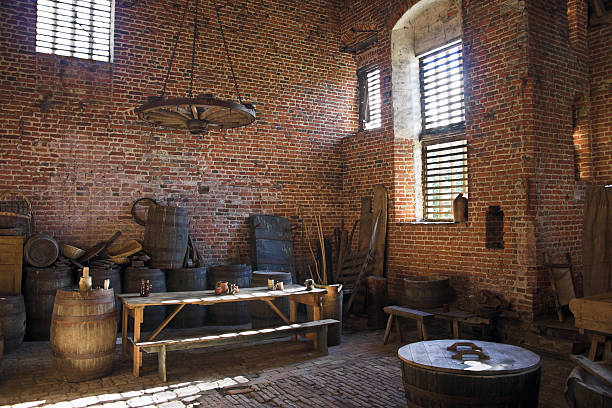 sunlight streams into an old victorian workman's room, containing tables,oak barrels,tools, used for workshop, storage, meals