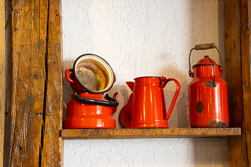 Old red kitchen pots on a wooden shelf in front of a white wall. Vintage metal pots and pitchers.