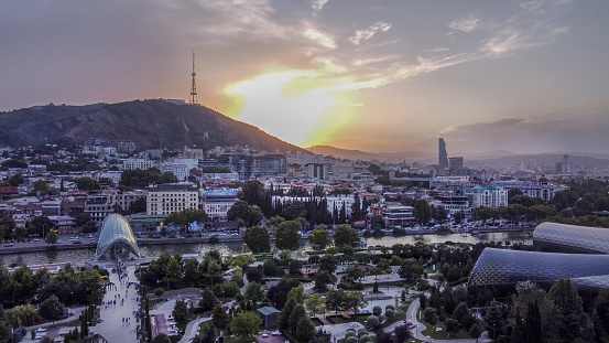 Fantastic  drone point of view on Tbilisi cityscape with  Kura river , pedestrian Bridge of Peace, Rike park  (people walking) , skyscraper (hotel?)  in right corner  and Hill with comminication tower on it   -  and orange sunset in sky