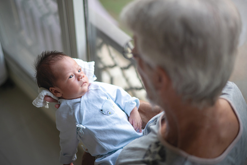 Senior man holding his one month old grandson at home during the day. Happy grandfather holding adorable newborn baby boy grandchild on arms by the window at home to expose to the daylight