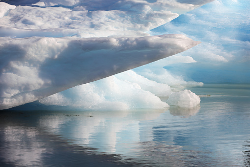 Part of a massive blue ice iceberg occupying the entire top two thirds of this horizontal frame.  Unusual pointed section of ice overhanging in the foreground.  The bottom third is its reflection in slightly rippled water.   Near Dawes Glacier.  Primary colors in the image are white, blues and gray-blue.  No people in the image.