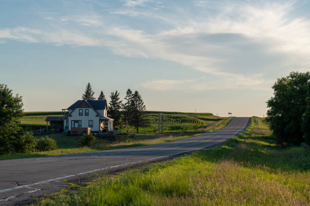 Looking down a country road lined with fields and a white farmhouse  on a calm evening.