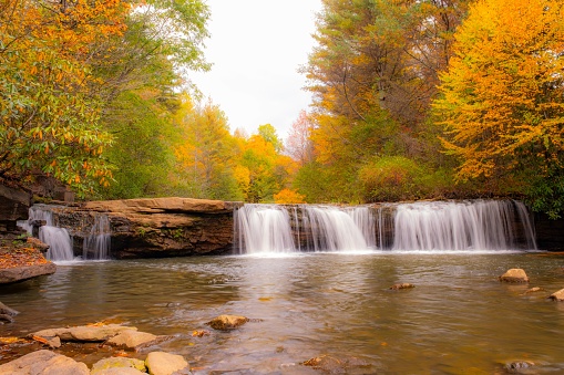 Long exposure photograph of waterfalls taken on a sunny fall day. The historic Pakenham five stone arched bridge is in the background, surrounded by trees showing fall colours.