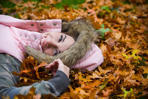 Cute young woman in hooded jacket looks at camera as she lays on a bed of Fall leaves. Copy space. CLICK FOR SIMILAR IMAGES AND LIGHTBOXES WITH MORE SEASONAL IMAGES OR BEAUTIFUL WOMEN. 