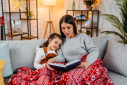 Smiling mother reading a storybook to her little daughter, curled up on the sofa under a soft blanket for Christmas.
