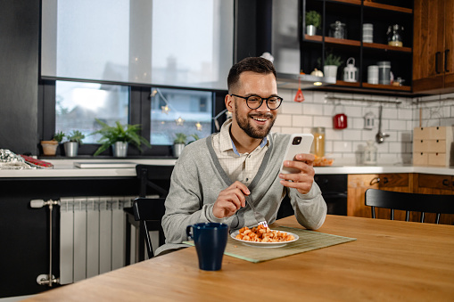 Portrait of a cheerful man sitting at the dining table and text messaging while eating
