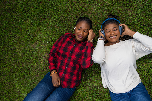 Two young black women lying on grass and listening music on headphones together.