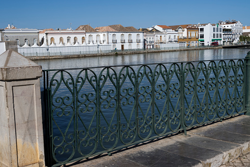 Decorative wrought iron railings in Tavira with the Gilao river and typical Algarve buildings in the background. Tavira is a city in the Algarve Province of Portugal and because it does not have an easily accessible beach it is less popular with the mass tourist trade and has retained much of its typical Algarvian character.