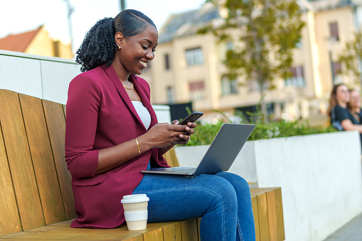 On a public bench in the city, an African American businesswoman creates her own work oasis, engrossed in her laptop tasks, a testament to her dedication