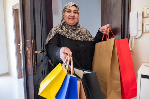 mulher árabe do oriente médio da jordânia segurando sacolas de compras e entrando em casa com sorriso no rosto usando hijab e abaya - women holding shopping bag living room - fotografias e filmes do acervo