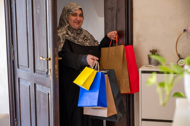 mulher árabe do oriente médio da jordânia segurando sacolas de compras e entrando em casa com sorriso no rosto usando hijab e abaya - women holding shopping bag living room - fotografias e filmes do acervo