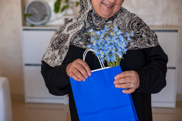 mulher árabe usando abaya e lenço de cabeça segurando saco de compras azul dentro da sala de estar com sorriso no rosto - women holding shopping bag living room - fotografias e filmes do acervo