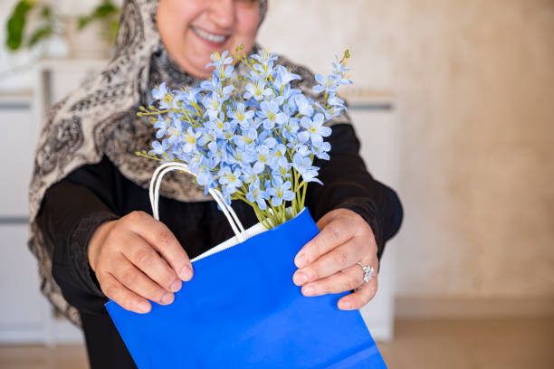 mulher árabe usando abaya e lenço de cabeça segurando saco de compras azul dentro da sala de estar com sorriso no rosto - women holding shopping bag living room - fotografias e filmes do acervo