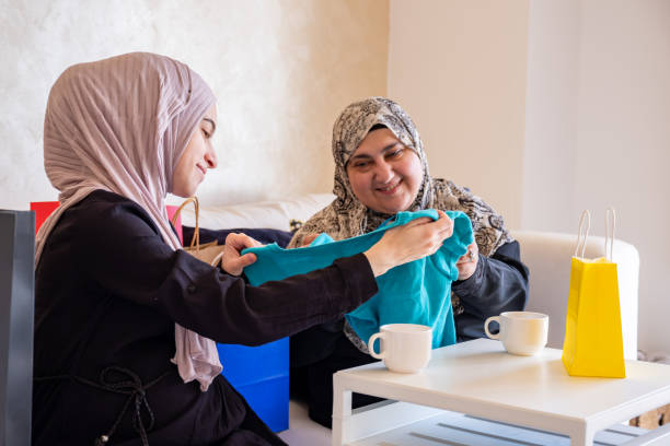 mulher árabe presenteando sua mãe e mostrando as roupas novas depois de fazer compras com sacolas de compras ao fundo na sala de estar - women holding shopping bag living room - fotografias e filmes do acervo